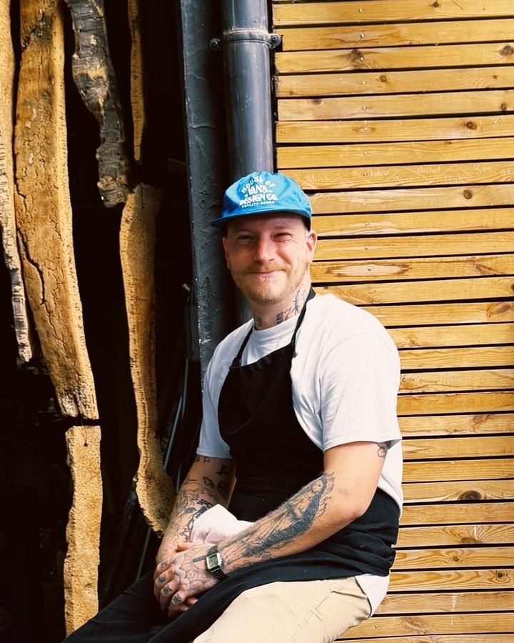 Portrait du chef à mi-corps, assis devant un mur de bois clair : casquette bleue, tablier noir, bras tatoués, mains jointes.
