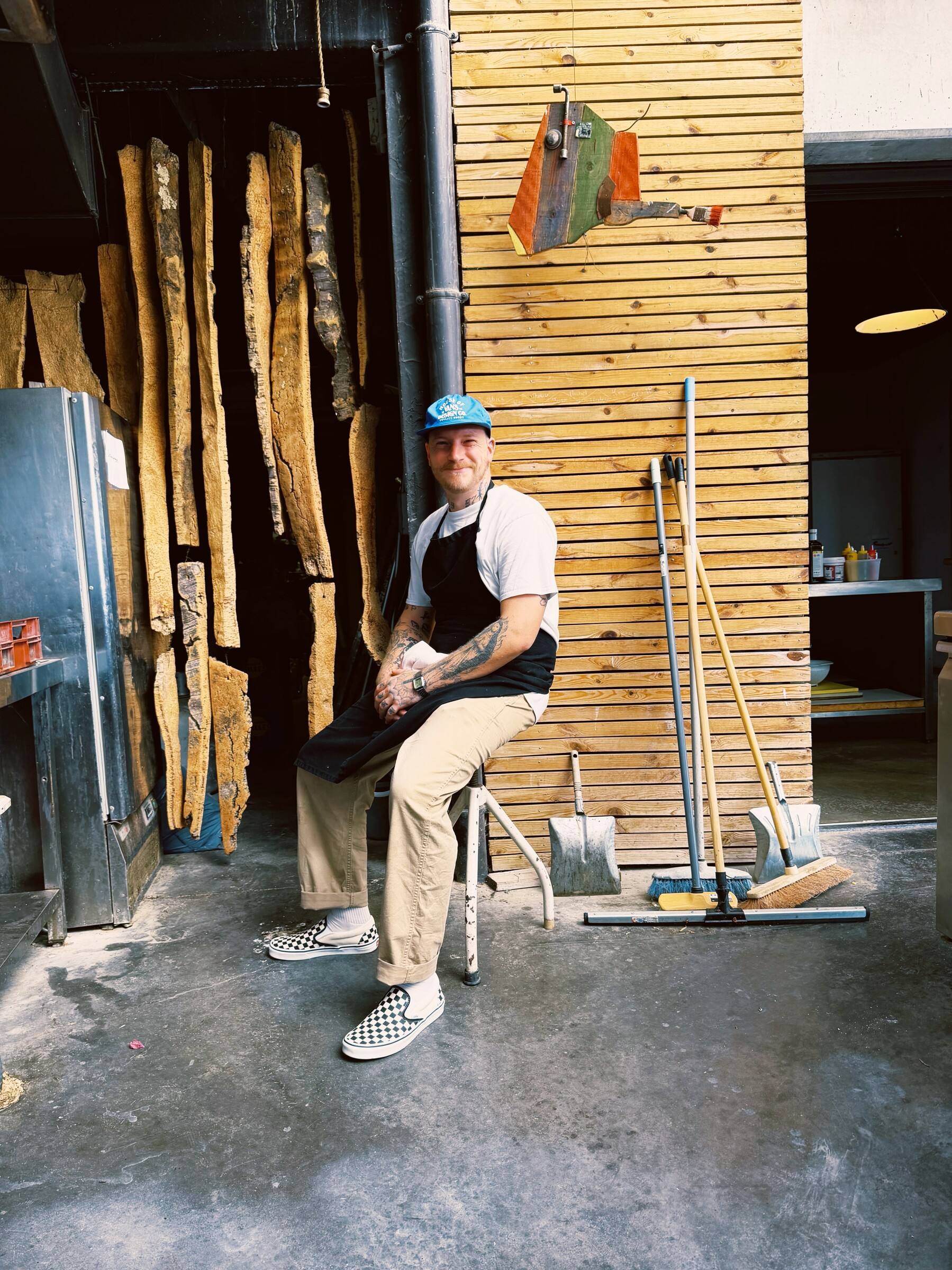 Le chef assis devant un mur de bois, casquette et tablier noir.