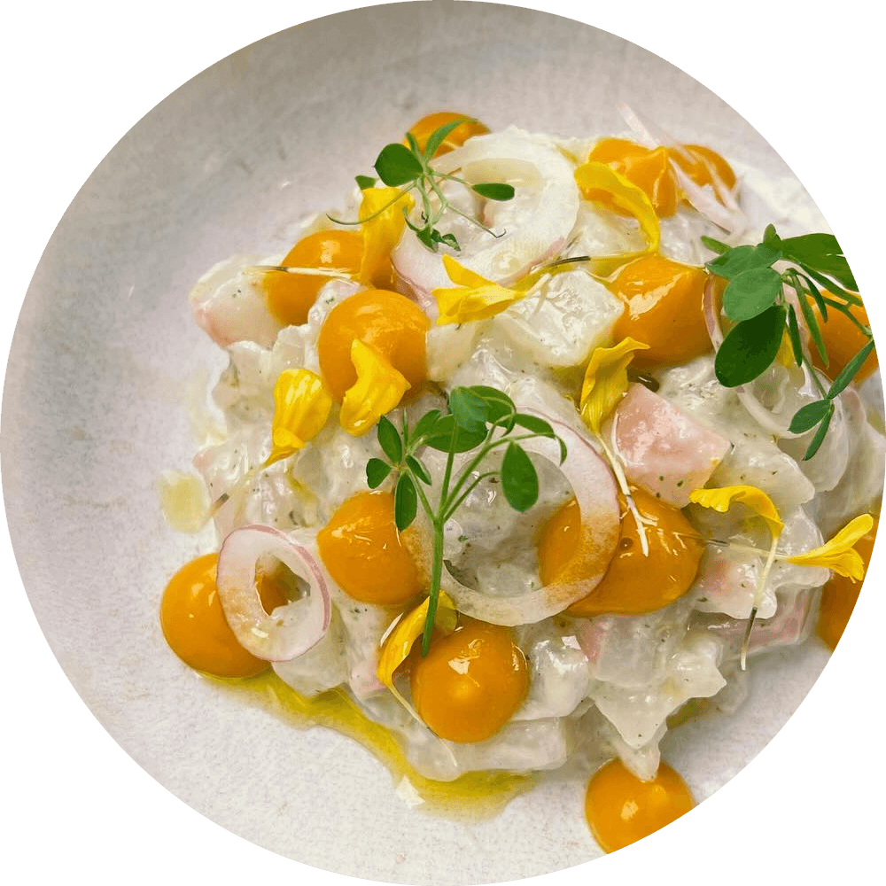 Crudo aux agrumes dans une assiette blanche, vu du dessus : points de gel orange, fleurs jaunes et jeunes pousses, en lune sur le ciel de nuit.