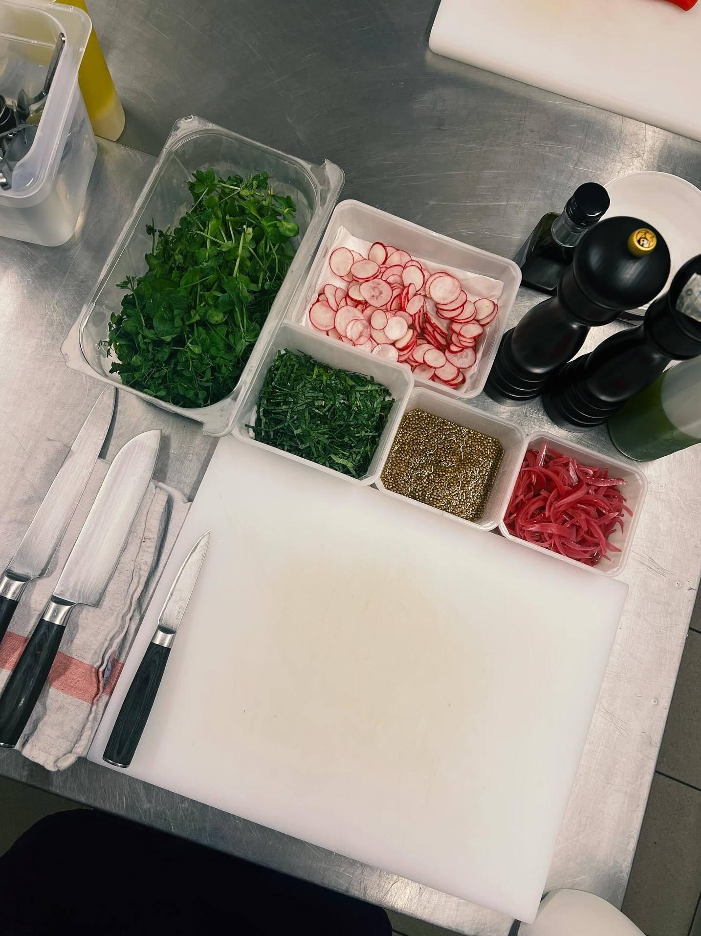 Mise en place en cuisine, vue du dessus : jeunes pousses, radis en fines rondelles, herbes ciselées, graines et lamelles d’oignon rouge en bacs, à côté des moulins à poivre et des couteaux.