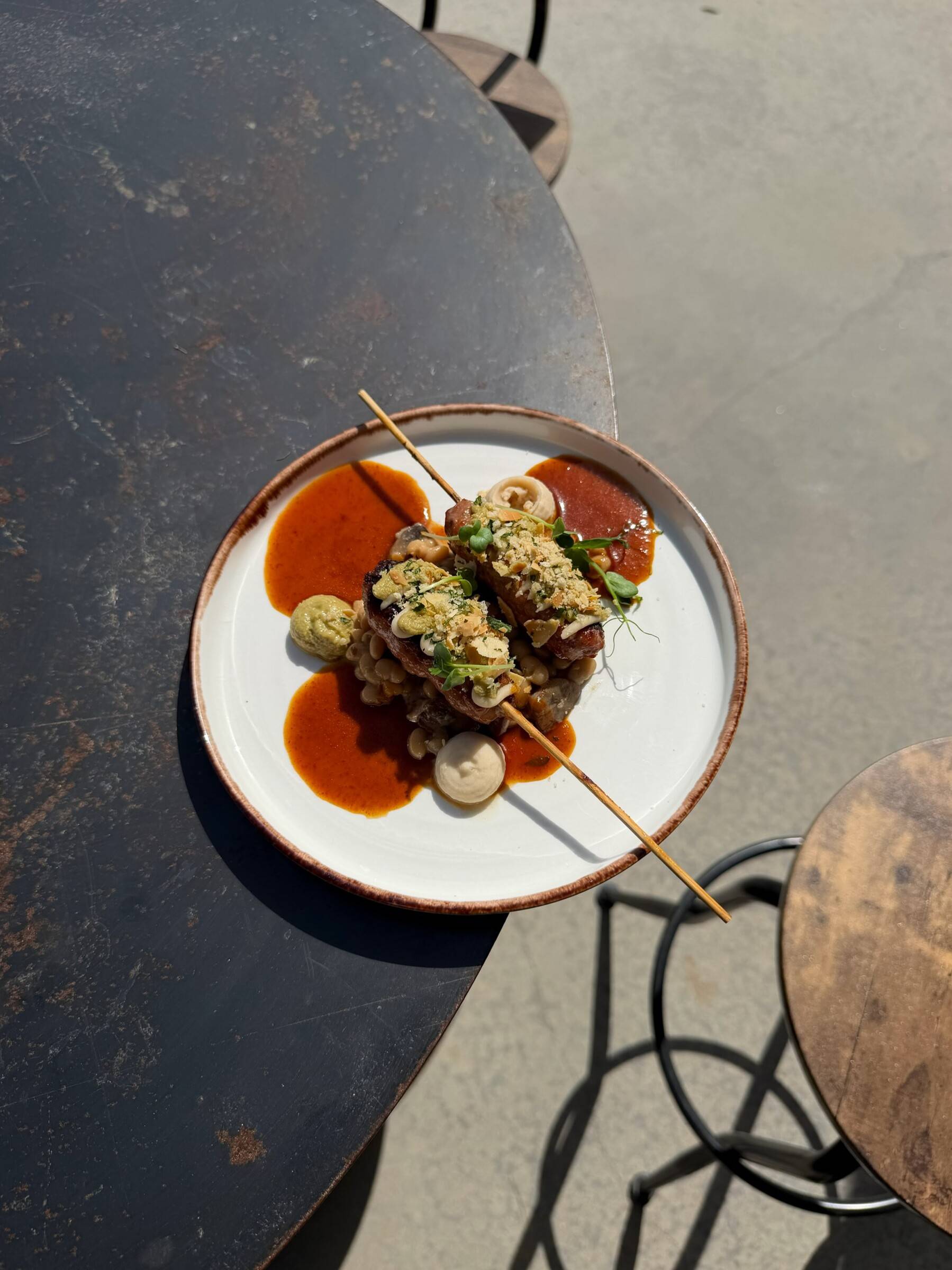 Brochette vue du dessus sur une assiette blanche, sauce rouge et condiment, sur une table en métal au soleil.
