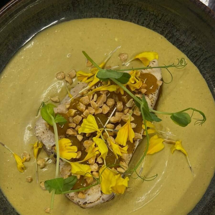 Sashimi de thon rouge dans un bouillon jaune, cacahuètes concassées, fleurs jaunes et jeunes pousses, dans une assiette creuse sombre.