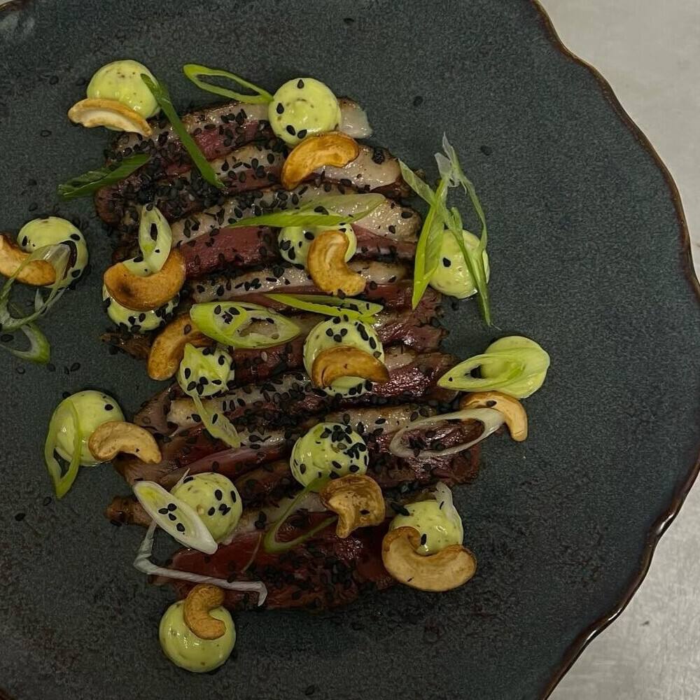Tranches de magret, noix de cajou torréfiées et points de mayonnaise verte, sur une assiette sombre, vue du dessus.