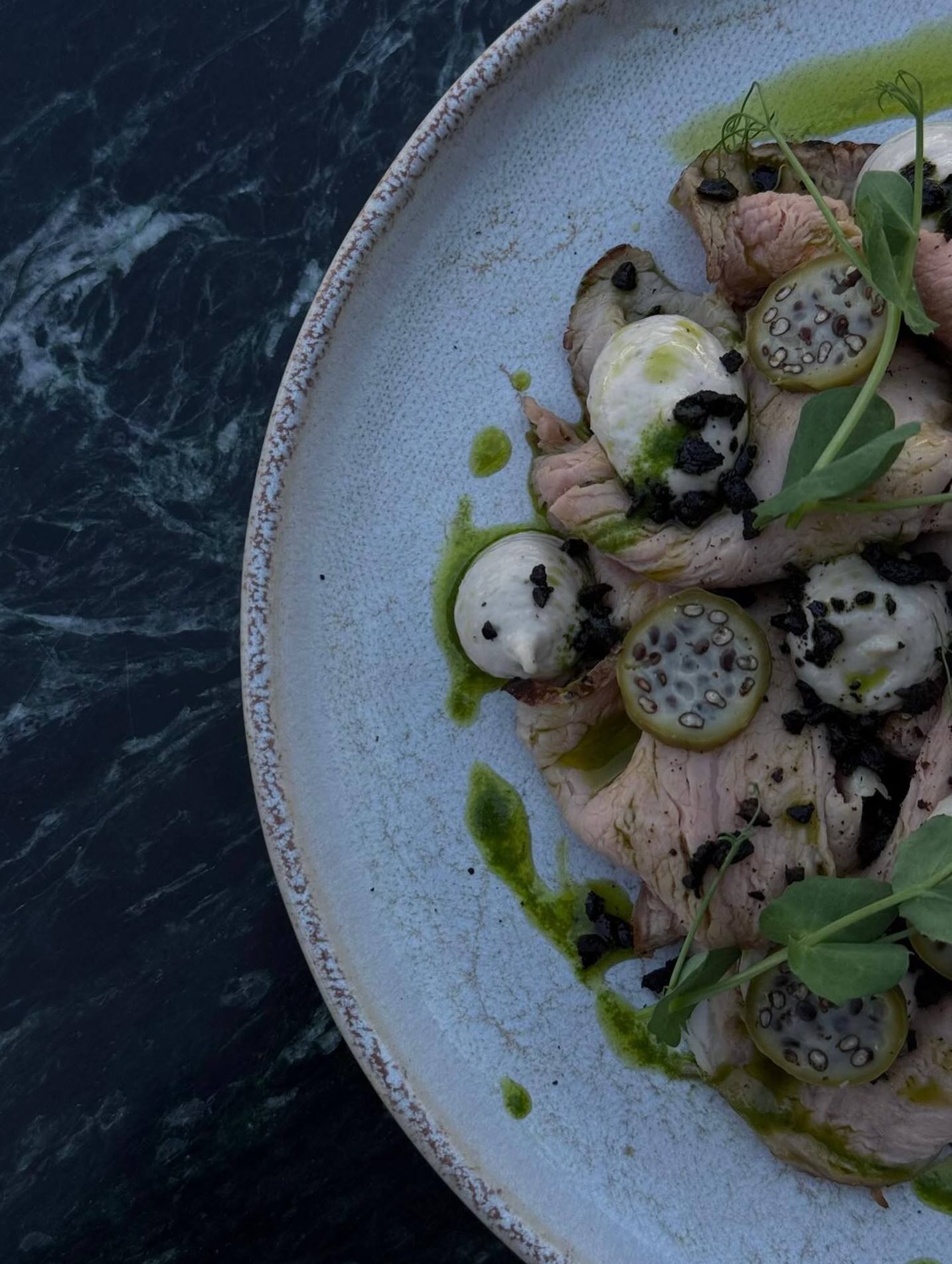 Détail du vitello tonnato dressé, vu du dessus : veau tranché en vagues, points de sauce, câpres à queue en rondelles, poudre d’olive, huile de roquette et pousses, sur une assiette en grès claire.