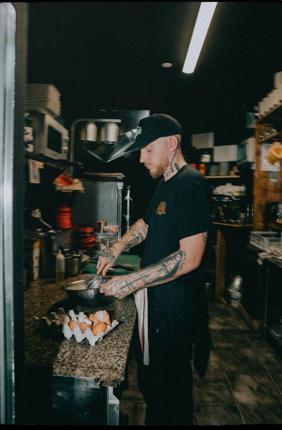 Le chef, casquette noire, cuisine au comptoir du Repère, une boîte d’œufs à côté de lui.