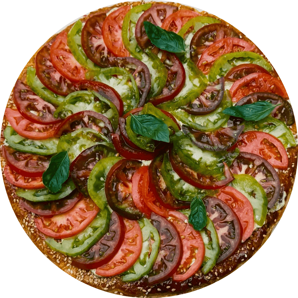 Tarte fine aux tomates vue du dessus, en soleil couchant : tranches de tomates rouges, vertes et noires en rosace, feuilles de basilic, bord de pâte doré au sésame.